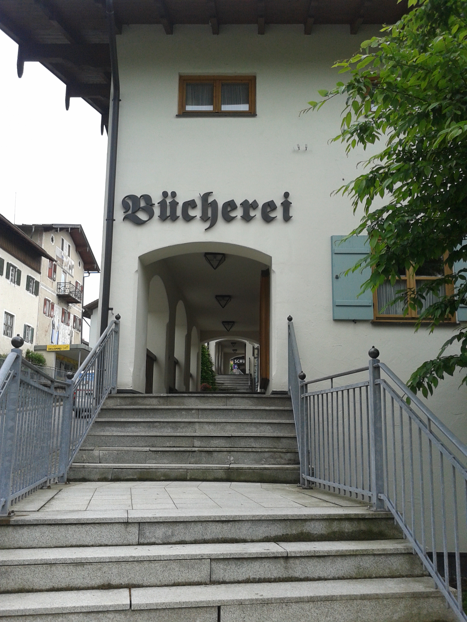 Eine Frontansicht der Bibliothek,vor dem Eingang eine Steintreppe mit Geländer. Die Treppe mündet in den Durchgang zu einer Fußgängerpassage. Am Eingang ein großer altdeutscher Schriftzug BUECHEREI.